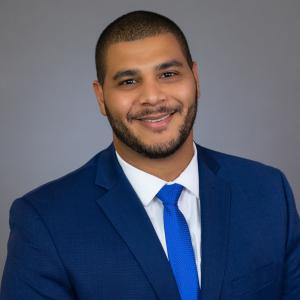 Jose Rodriguez in dark blue suit with bright blue tie with a gray white gradient background