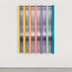 A modern wall sculpture featuring seven vertical bars in pastel colors—yellow, orange, coral, pink, lavender, light blue, and teal—mounted over a reflective metallic background with curved surfaces. The artwork is displayed against a white wall and gray floor.
