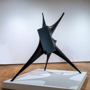 Abstract black metal sculpture with three pointed legs and a central geometric form, displayed on a white platform in a gallery.