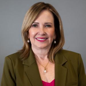 Martha Pantin headshot with olive blazer and light red shirt, gray gradient background