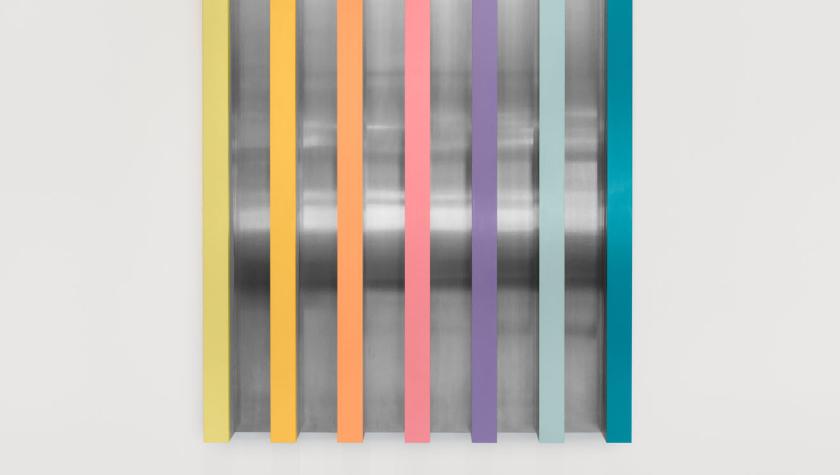 A modern wall sculpture featuring seven vertical bars in pastel colors—yellow, orange, coral, pink, lavender, light blue, and teal—mounted over a reflective metallic background with curved surfaces. The artwork is displayed against a white wall and gray floor.