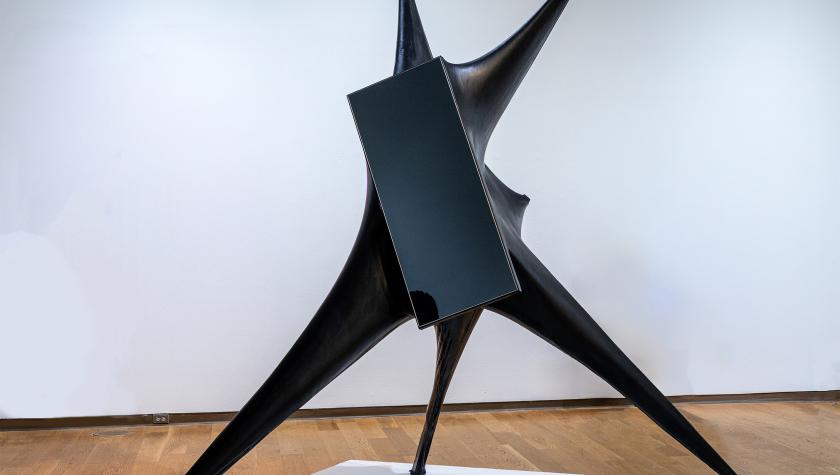 Abstract black metal sculpture with three pointed legs and a central geometric form, displayed on a white platform in a gallery.