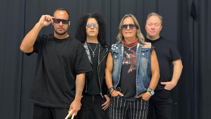Soul Travelers band in front of a black curtain, 4 dressed in black and rock-inspired attire; one holds drumsticks, another wears a leather jacket, one wears a denim vest and striped pants, and all face the camera with a confident, band-style pose.