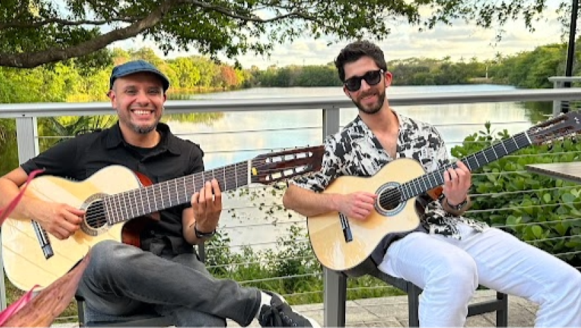 Two guitar players sitting outside smiling, behind them a lake view