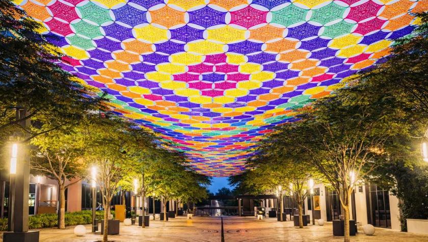 Colorful crocheted canopy in purple, yellow, orange, pink, and green stretches above  tree-lined pedestrian (Giralda Plaza) walkway illuminated at dusk.