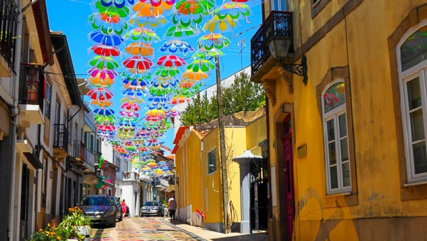 Colorful flower-pattern umbrellas hang above a narrow street lined with buildings, casting bright floral shadows across the cobblestone road.