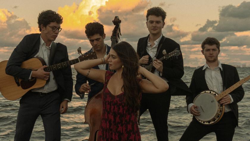 The Wire Jays posed photo, four men in a suits with instruments and a woman in front in a red dress. A yellow orange sun sets behind the clouds and ocean