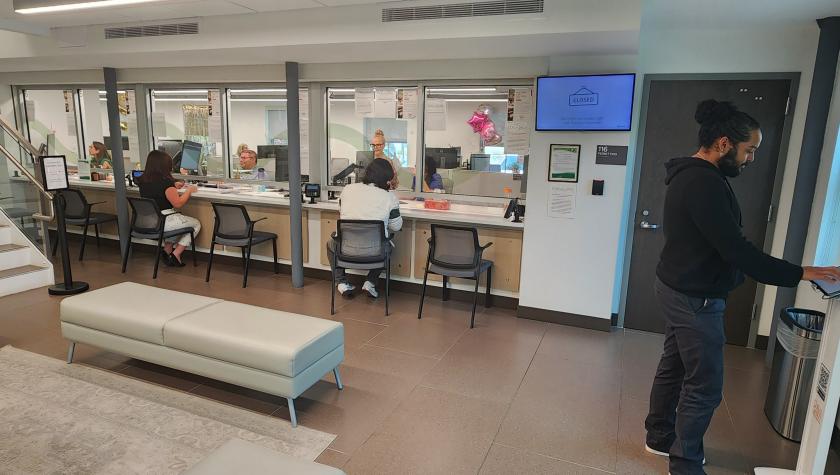 In waiting room area with help windows, customers talk to employees and a man in black sweater signs into an ipad