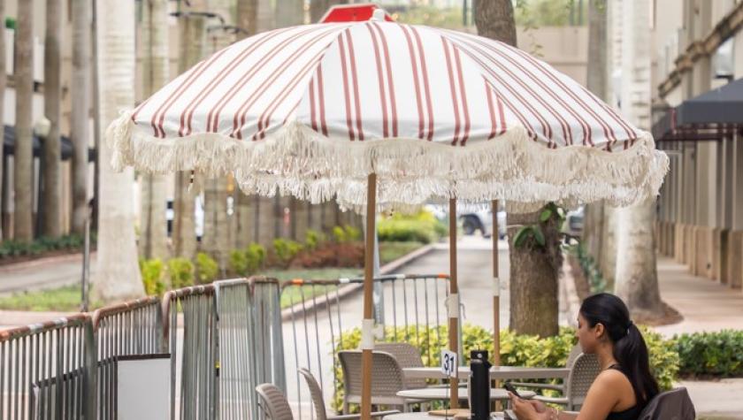 A woman sits on a chair under a line pattern umbrella on the street in a parking day lawn spot